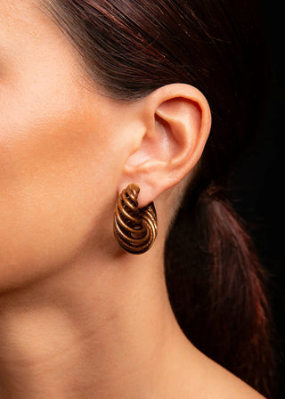 A close-up of a model's ear wearing a textured, twisted gold hoop earring against a dark background.