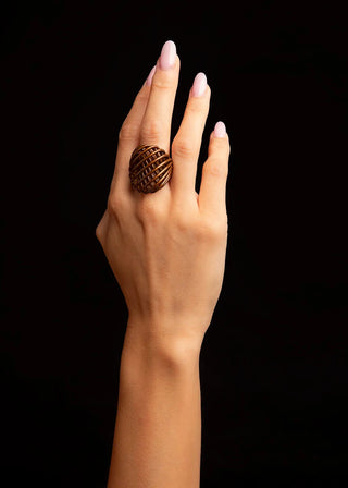 A close-up of a hand with pale pink manicured nails wearing a large, textured bronze ring, set against a solid black background.