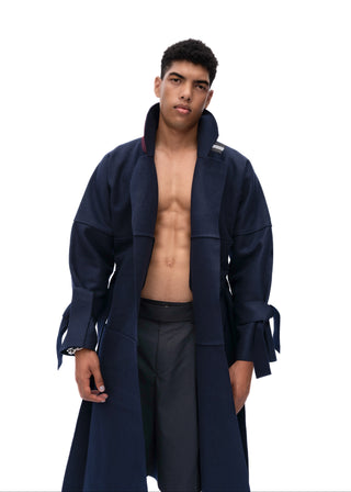 A male model wearing a long navy blue open-front coat over a bare torso and dark trousers, photographed against a plain white studio background.