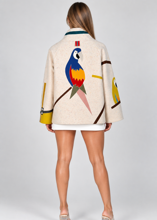 A model shown from the back wearing a cream-colored jacket with a large colorful parrot graphic on the back and smaller parrot graphics on the sleeves, paired with a white mini skirt and clear high-heeled sandals.
