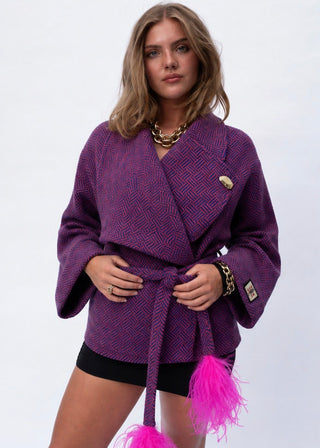 A woman models a purple textured wrap jacket with a matching tie belt, featuring bright pink feather accents at the belt ends and gold-tone hardware, set against a plain white studio background.