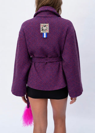 A woman shown from the back wearing a purple and red patterned belted jacket with a small square patch on the upper back, paired with a black skirt. She is holding a bright pink tassel accessory against a plain white studio background.