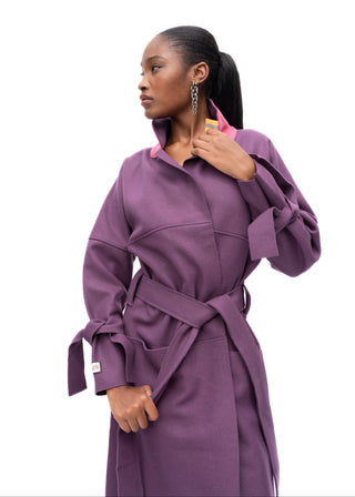 A woman modeling a purple belted coat with a pink collar detail, photographed against a solid white studio background.