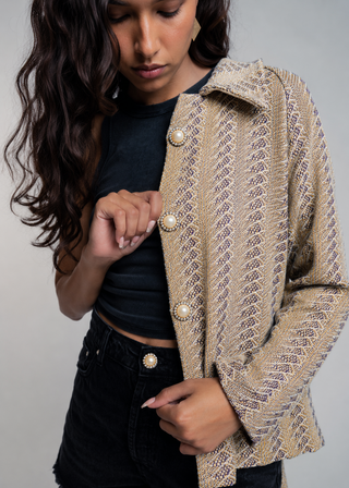 A woman wearing a beige textured tweed jacket with a geometric pattern, large pearl-like buttons, and a collar, styled over a black crop top and black denim shorts against a neutral gray background.
