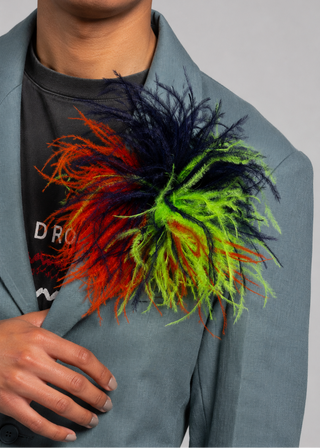 A close-up of a person wearing a large, vibrant, multi-colored feathered accessory on their lapel. The accessory features a mix of neon green, orange, red, and dark navy blue feathers.