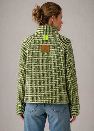 A woman viewed from the back wearing a green and white patterned zip-up jacket with a brown leather patch on the back, paired with blue jeans, against a solid gray studio background.