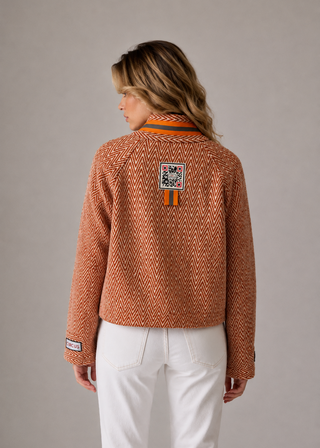 A woman viewed from the back wearing a cropped, textured herringbone knit sweater in a rust-orange color with white pants, set against a plain gray studio background.