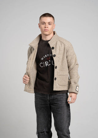 Man wearing a beige jacket over a black t-shirt with visible branding on a gray background