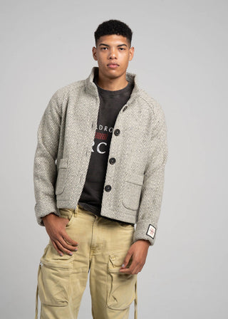 Man wearing a textured beige jacket over a black graphic t-shirt with beige pants on a gray background