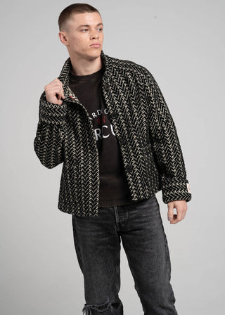 Man wearing a patterned jacket over a black t-shirt with visible branding on a gray background