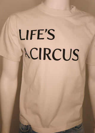 A cream-colored short-sleeve t-shirt with the text 'LIFE'S A CIRCUS' printed in black across the chest, displayed on a mannequin torso with blue jeans visible at the bottom.