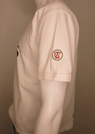 A mannequin wearing a light beige short-sleeve t-shirt, shown from a side profile. The shirt features a small circular red and black logo on the left sleeve and a partial black graphic on the right chest.