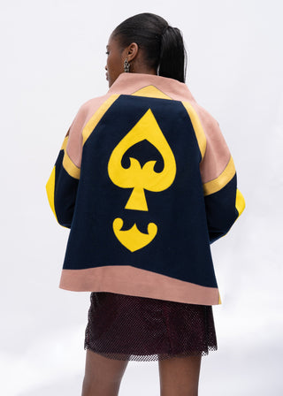 Woman wearing a navy and yellow wool jacket with a large yellow spade symbol on the back, standing against a white background.