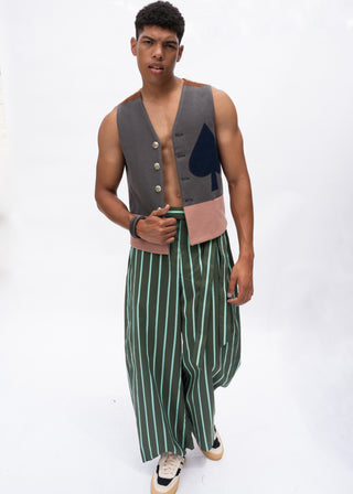 A male model wearing a unique two-piece outfit consisting of a gray vest with a large black spade graphic and a dusty pink waistband, paired with green and white vertically striped wide-leg pants, on a white studio background.
