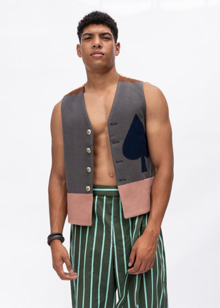 A man modeling a sleeveless vest with a gray front panel, pink side panels, gold buttons, and a dark blue spade graphic, paired with green and white striped trousers, against a plain white background.