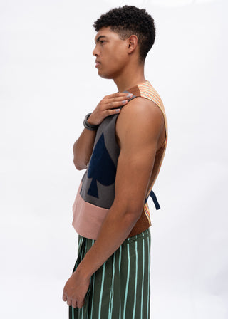 A man in profile wearing a sleeveless knit vest with geometric color-block panels in beige, gray, navy, and blush pink, paired with green and white vertically striped pants, against a plain white studio background.