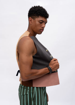 A man in profile wearing a dark gray sleeveless vest with gold buttons over a striped orange and white top, paired with green and white striped trousers, holding a dusty pink folder against a light gray background.