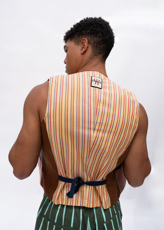 A man viewed from behind wearing a sleeveless striped vest with orange, yellow, and blue vertical stripes, a dark brown waist tie, and green striped shorts against a light gray background.