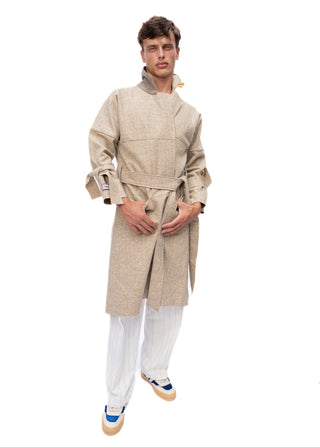 A young man modeling a beige textured long coat with a tie belt, paired with white pinstriped trousers and blue sneakers, against a solid white background.