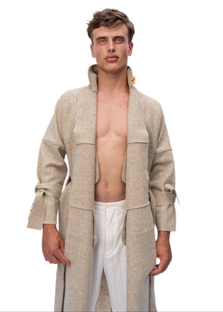 A male model wearing a long, open-front beige wool coat over a white shirt and white trousers, standing against a solid white background.