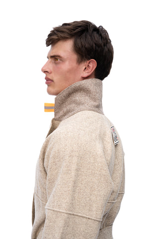 A man in profile wearing a beige textured high-collar jacket against a solid white background.