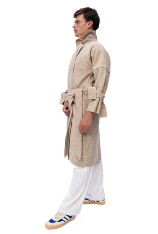A man shown in profile wearing a beige textured belted coat over white pleated trousers and blue sneakers, isolated on a white background.