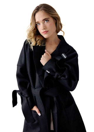 A woman wearing a black belted coat with a shawl collar, photographed against a solid white background.