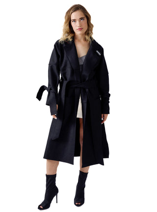 A woman modeling a long black wrap-style coat with a tie belt, shown against a plain white studio background. She is wearing black ankle boots and a gray and white outfit underneath.