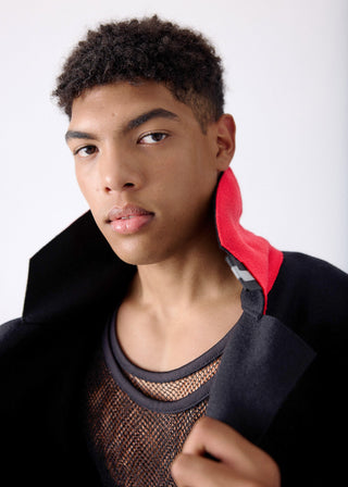 A close-up portrait of a young man wearing a black coat over a brown mesh top with a bright red inner collar, set against a plain light gray background.