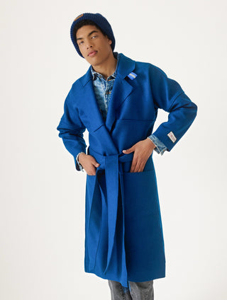 A model wearing a bright blue wool-blend coat with a matching waist tie belt, styled over a denim shirt and paired with a blue knit beanie, photographed against a clean white studio background.