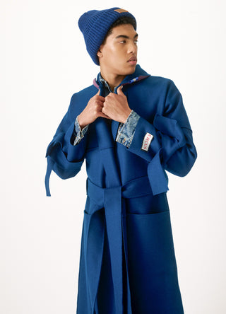 A model wearing a long, oversized royal blue coat with a matching belt, a blue knit beanie, and a denim shirt underneath, posed against a solid white studio background.