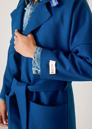 A close-up of a person wearing a bright blue belted coat over a light blue denim shirt. The coat has a visible white rectangular patch on the cuff with the word 'CIRCUS' in red text.