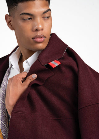 A close-up of a man wearing a maroon wool-blend blazer over a white collared shirt and patterned tie, holding the lapel of the blazer against a plain white background.