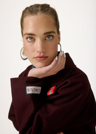 A close-up portrait of a woman wearing a burgundy fleece pullover with a circular patch reading 'CIRCUS' on the sleeve and a small red-and-white striped detail on the chest, against a plain light background.