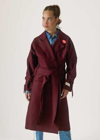 A woman modeling a long maroon belted coat over a blue denim shirt and jeans, photographed against a plain light gray studio background.
