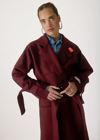 A model wearing a long, deep burgundy wrap coat with a tie waist, styled over a blue denim shirt. The coat features a structured collar, wide lapels, and a matching belt, with a small red tag on the chest pocket.