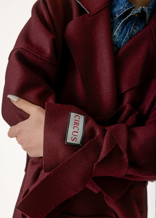 A close-up of a person wearing an oversized burgundy coat with a matching tie belt and a visible rectangular patch reading CIRCUS. A blue denim shirt is partially visible underneath.