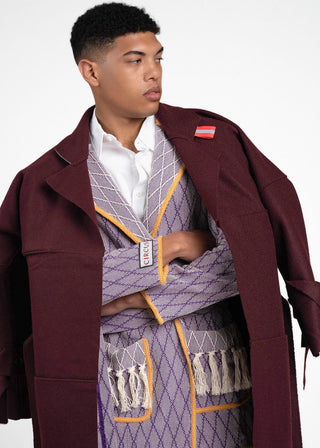A model wearing a burgundy oversized coat over a purple and white patterned robe with yellow trim, layered over a white collared shirt, against a plain white studio background.