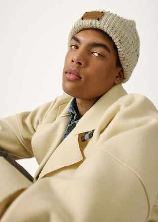 A man wearing a cream-colored wool coat, a beige knit beanie with a brown leather patch, and a blue patterned shirt, photographed against a clean white background.