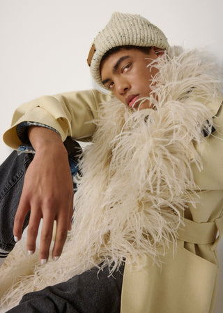 A model wearing a beige trench coat with a large white feathered collar, a cream knit beanie, and dark jeans, posed against a plain off-white background.
