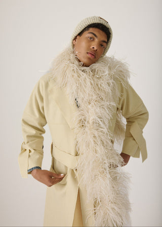 A model wearing a cream-colored coat with a large fluffy feathered collar, a matching knit beanie, and a long cream feather boa, posed against a plain light gray studio background.