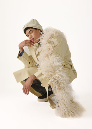 A model crouches in a studio setting wearing a beige trench coat with a voluminous cream feathered collar, a cream knit beanie, and dark pants against a plain white background.