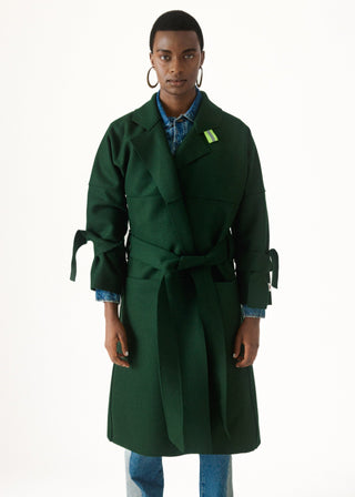 A model wearing a dark forest green belted coat over a blue patterned shirt and jeans, standing against a plain white studio background.
