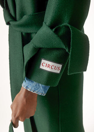Close-up of a dark green wool coat with a matching belt, featuring a cuff patch with the brand name 'CIRCUS' in red text on a white background, worn over a light blue denim shirt.