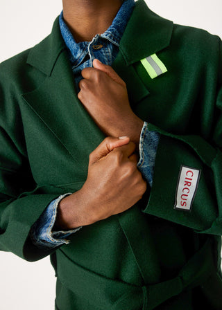 A close-up of a person wearing a dark green jacket with a white patch reading 'CIRCUS' on the cuff, layered over a blue denim shirt, against a plain light background.
