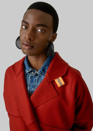 A model wearing a vibrant red wool-blend coat over a blue denim shirt, featuring a small orange and gray striped lapel pin, set against a plain light gray background.