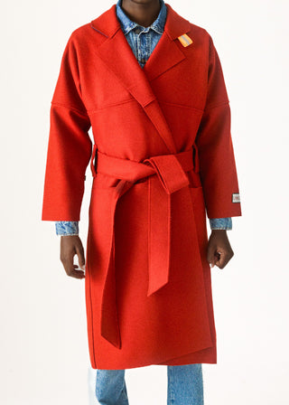 A model wearing a bright red wool wrap coat with a matching waist tie belt, layered over a blue denim shirt and jeans, against a plain white background.