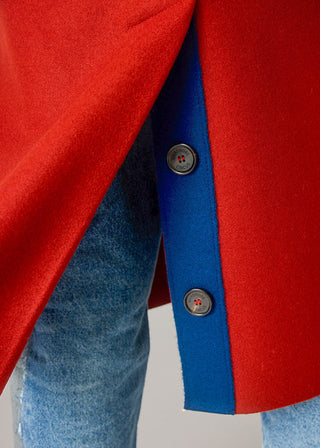 Close-up of a red coat with a bold blue vertical stripe and dark buttons, worn over blue jeans.