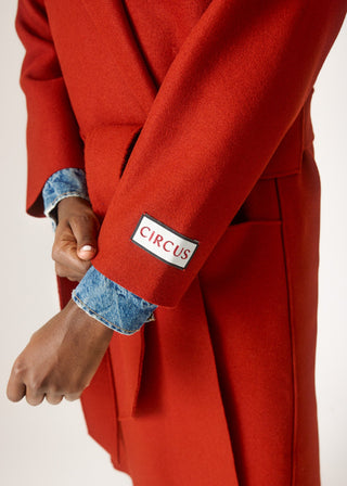 A close-up of a person wearing a bright red wool coat with a white rectangular patch on the cuff that reads 'CIRCUS' in red letters. The person is also wearing a blue denim shirt underneath, and the background is a clean off-white studio setting.