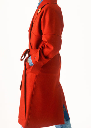 A side profile view of a person wearing a long, bright red wool coat with a tie belt, layered over a blue patterned shirt and light blue jeans, set against a plain white background.
